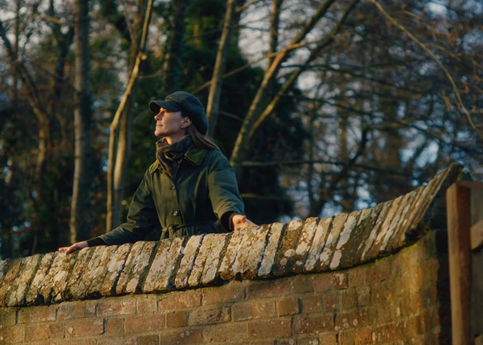 Woman in green jacket and cap standing behind a brick wall, reflecting outdoors, related to Kate Middleton and Meghan Markle. Woman in green jacket and cap standing behind a brick wall, reflecting outdoors, related to Kate Middleton and Meghan Markle.