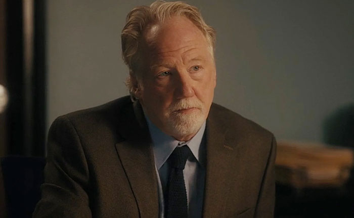 Middle-aged man with gray hair and beard wearing a suit in a serious scene related to US Marshals manhunt for Timothy Busfield.