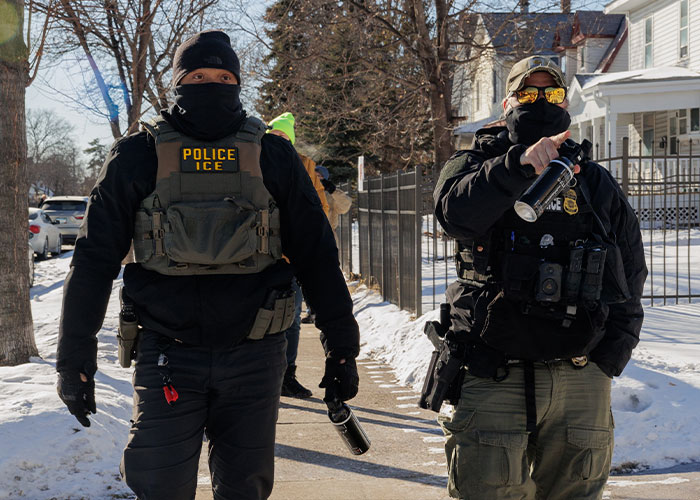 Two ICE officers dressed in tactical gear holding pepper spray while walking on a snowy residential sidewalk. Two ICE officers dressed in tactical gear holding pepper spray while walking on a snowy residential sidewalk.