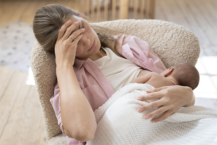 New mom holding baby, looking stressed and tired while sitting in a chair, reflecting challenges with husband and in-laws.