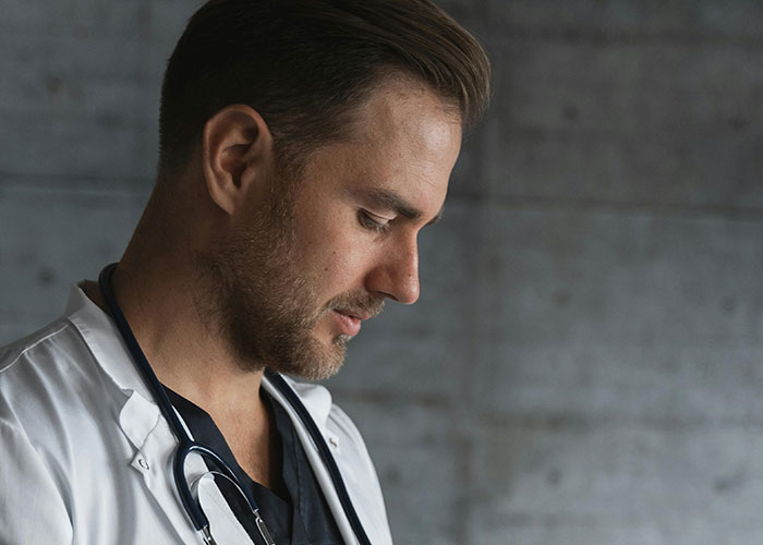 Young male medical professional with stethoscope, looking down thoughtfully in a clinical setting about patient care challenges.