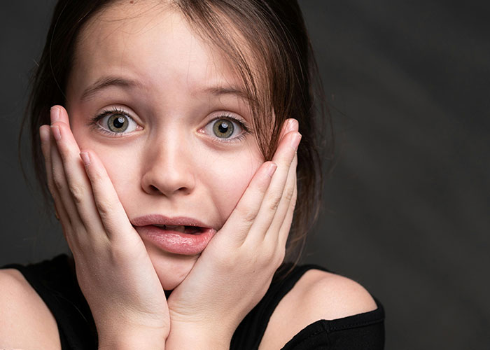 Young girl with rare physiological traits looks surprised, holding her face in her hands against a dark background.