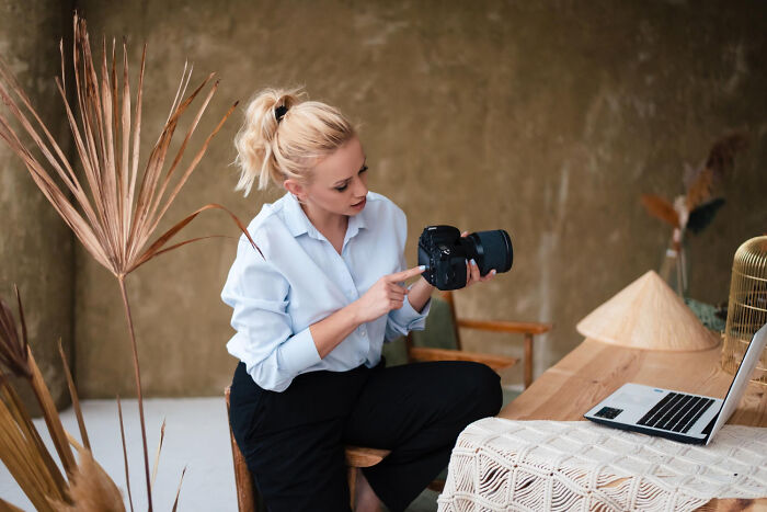 Young woman inspecting camera while sitting at table with laptop, illustrating satisfying situations and not my problem moments.
