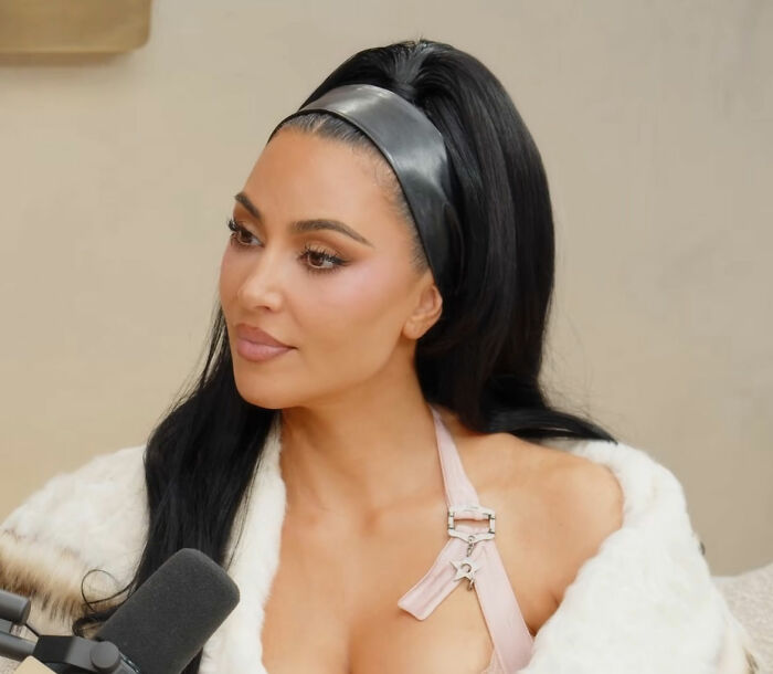 Kim Kardashian speaking during an interview, wearing a white fur coat and a black headband with sleek dark hair.