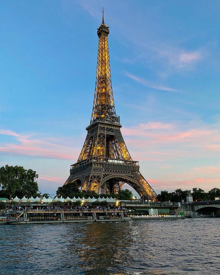Eiffel Tower lit at dusk with river and boats in foreground, illustrating countries that sleep the most concept.