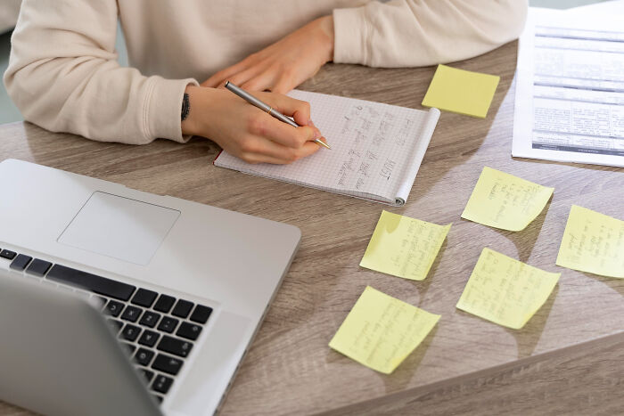 A person writing notes on a notepad at a desk with a laptop and multiple sticky notes showing bizarre office rules.