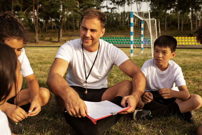 Coach sitting on grass with children during soccer practice, illustrating weird and satisfying situations not their problem anymore.