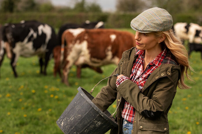 Woman in a plaid shirt and cap holding a bucket near cows in a field, illustrating fastest times couples said I do and done.