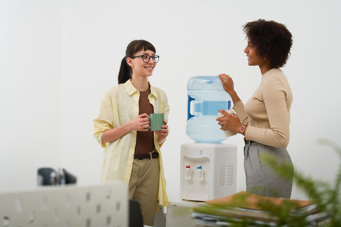Two colleagues chatting and drinking water by the office water cooler, illustrating bizarre office rules.
