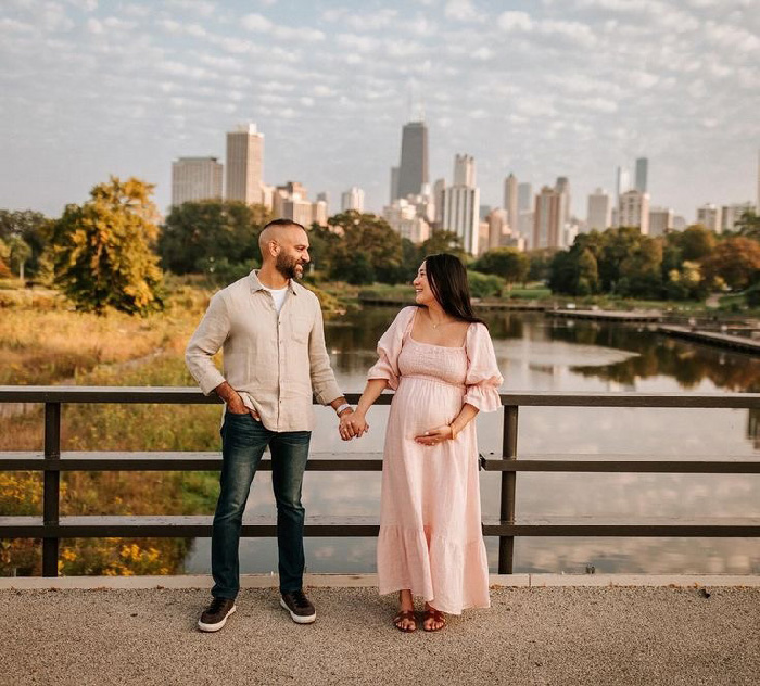 Couple holding hands on a bridge by a lake with city skyline, pregnant woman in pink dress, maternity photoshoot