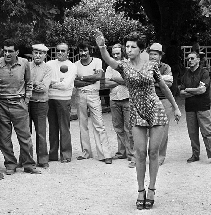 Woman in a patterned dress playing ball game outdoors, surrounded by men, capturing a moment in history of humanity.
