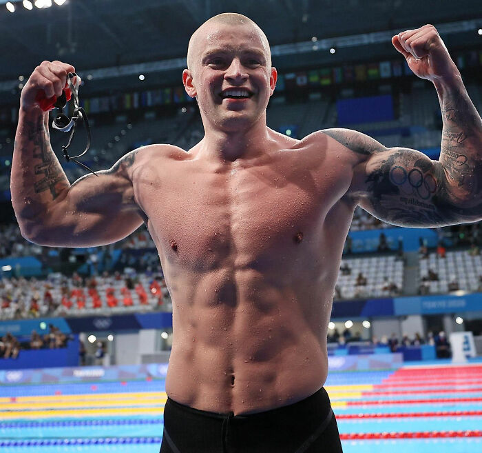 Adam Peaty celebrating his swimming victory, flexing muscles and holding goggles at an indoor aquatic center.