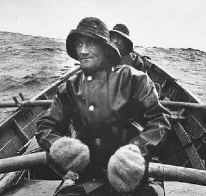 Fishermen wearing heavy coats and mittens rowing a boat in rough seas in a historical photograph of humanity.