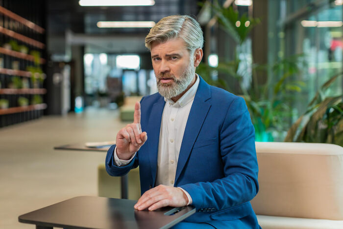Middle-aged man in blue suit gesturing with finger, conveying a weird and iconic not my problem situation.