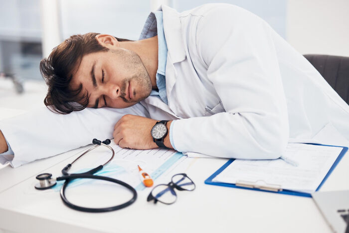 Doctor sleeping on desk showing unethical behavior with patient documents and stethoscope nearby in clinical setting.