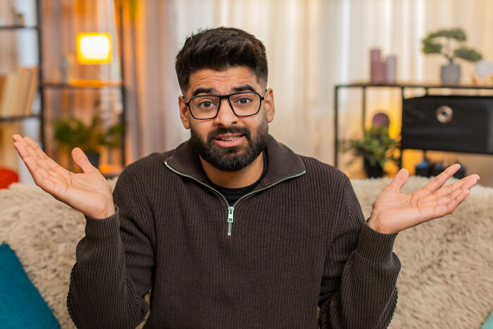 Man shrugging with raised hands in cozy room, capturing weird iconic satisfying situations not my problem anymore feeling.