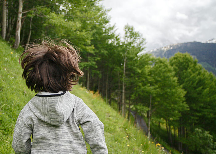 Child with messy hair wearing a gray hoodie running on a grassy hill surrounded by trees, capturing wacky stories outdoors.