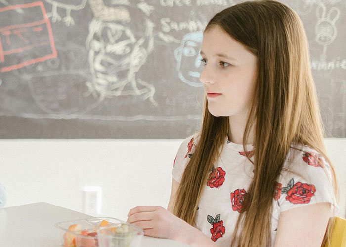 Young girl sitting at table with bowl of fruit, sharing family lore stories in a classroom setting for kids with no filter.