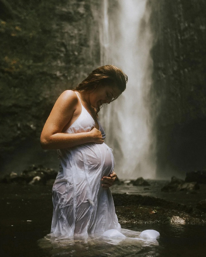 Pregnant woman in wet white dress holding belly during maternity photoshoot at a waterfall, standing in shallow water