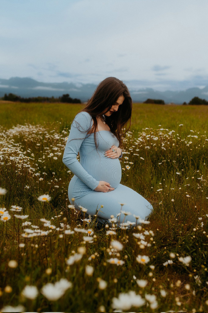 Pregnant woman in blue dress kneeling in a wildflower field, hands on belly, creative maternity photoshoot.