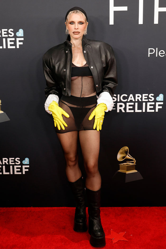 Bold and wild Grammys look featuring black sheer dress, leather jacket, yellow gloves, and knee-high boots on red carpet.