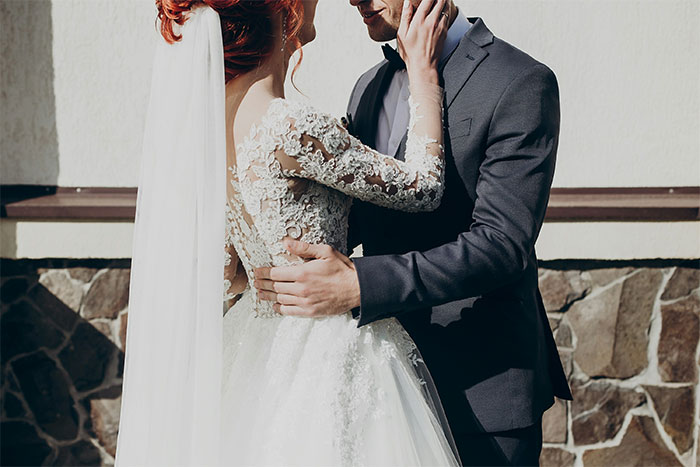 Bride in lace wedding dress and veil embracing groom in dark suit outdoors near stone wall background