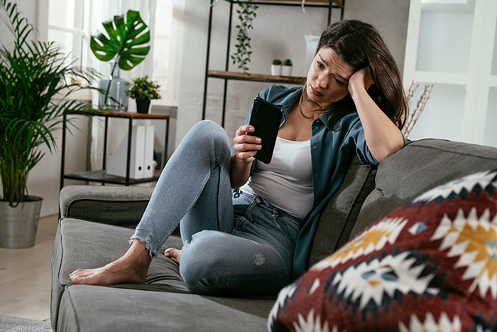 Woman sitting on couch looking upset at phone, reflecting on karma after affair partner leaves her alone.