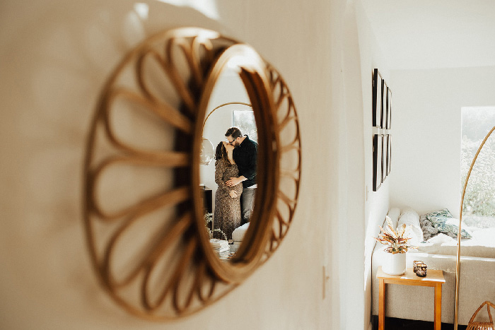 Pregnant couple kissing reflected in decorative round mirror, cozy living room maternity photoshoot ideas