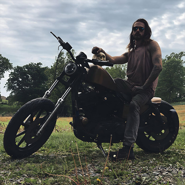 Man sitting on a motorcycle holding a skull outdoors with trees in the background, suggesting a chilling discovery.