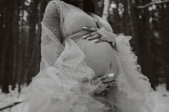 Black-and-white maternity photoshoot of pregnant woman in flowing tulle gown, tattooed hands cradling belly in snowy forest