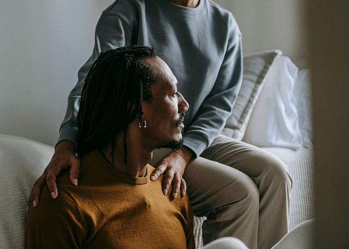 Two people indoors, one sitting on couch with supportive hand on shoulder, illustrating neurodivergent habits and understanding.