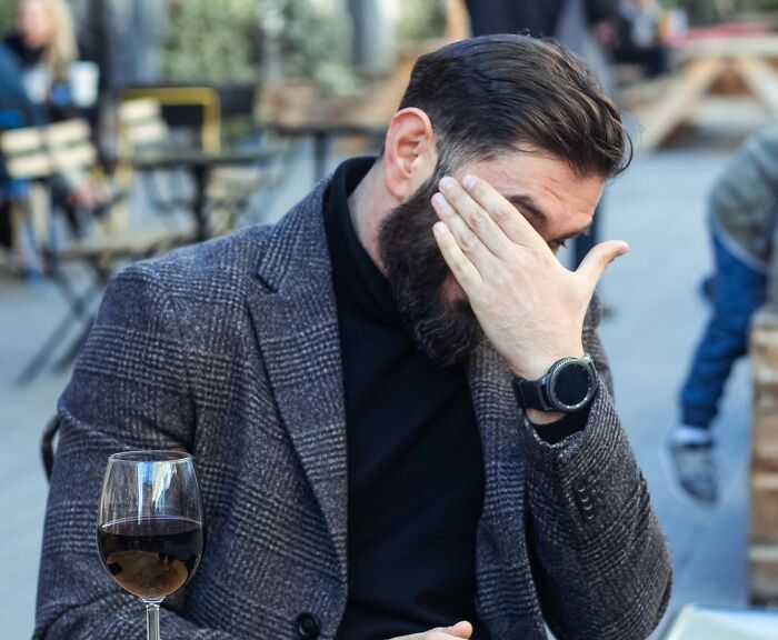 Man in a blazer sitting outdoors with a glass of red wine, covering his face in an awkward moment in the service industry.