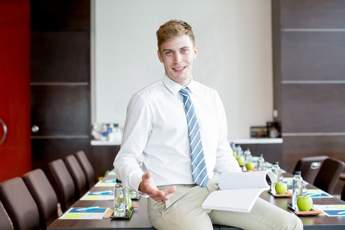 Young professional in a white shirt and striped tie explaining bizarre office rules in a modern conference room setting.