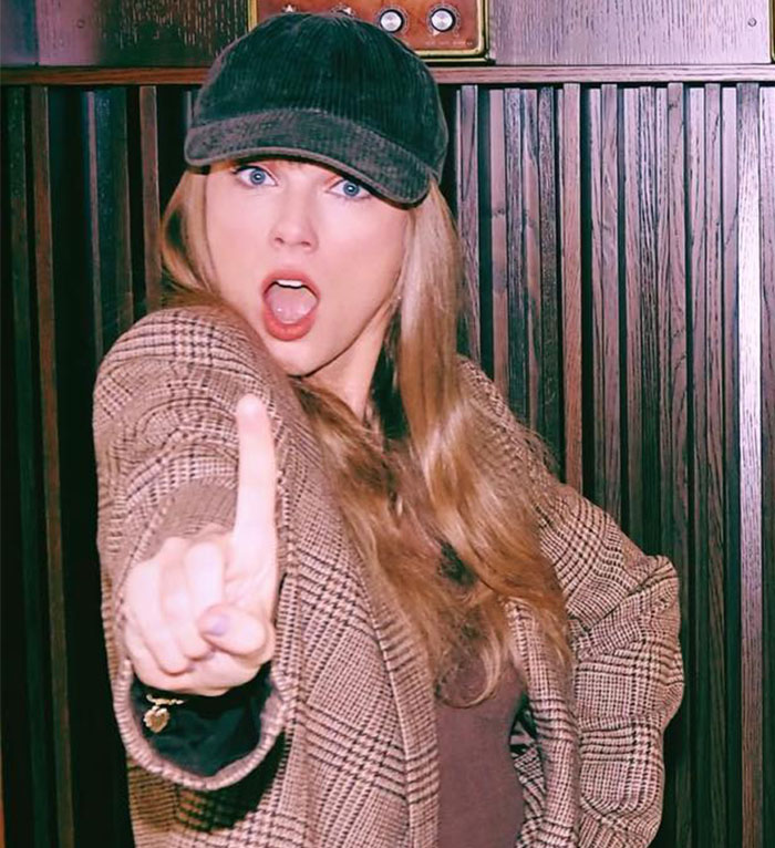 Taylor Swift wearing a cap and plaid jacket, making a bold gesture with an intense facial expression indoors. Taylor Swift wearing a cap and plaid jacket, making a bold gesture with an intense facial expression indoors.