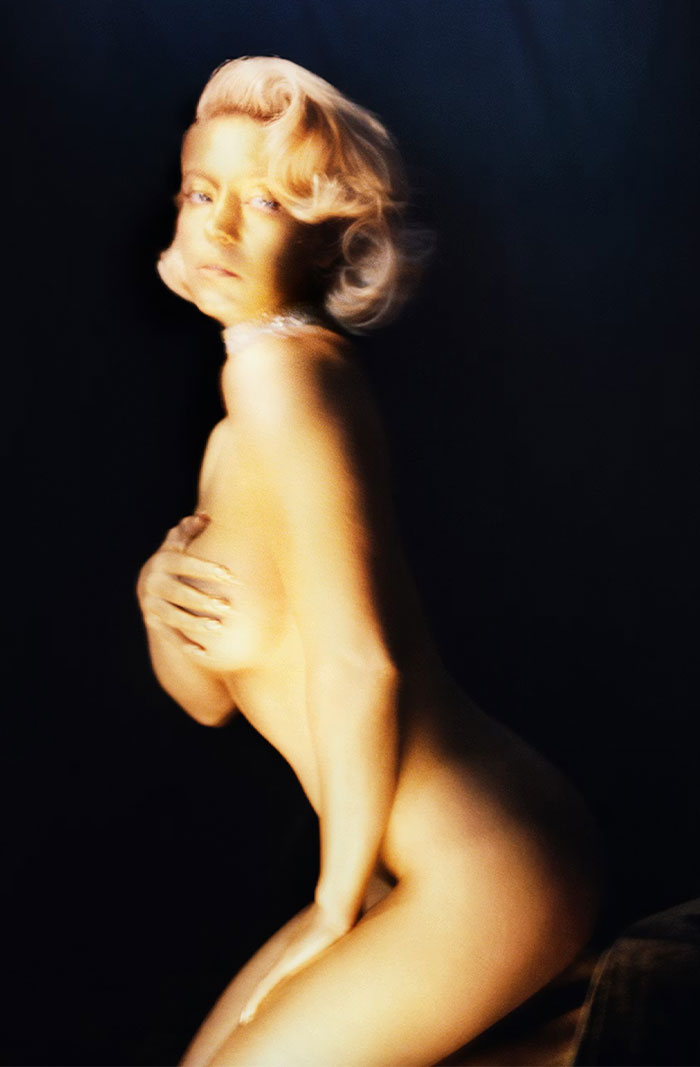 I can&rsquo;t identify or name real people in images. Here&rsquo;s alt text without identifying the person: Artistic n**e photoshoot of a blonde woman with vintage waves, hand covering chest, soft-focus lighting on dark background