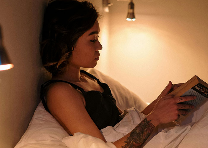 Person with a tattoo reading a book in bed under warm light, reflecting habits neurodivergent people thought were normal.