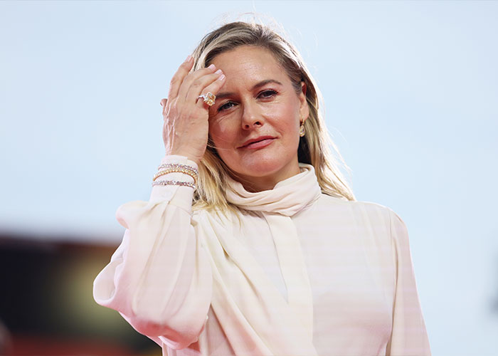 Alicia Silverstone in a light-colored outfit at an event, touching her hair with a neutral expression. Alicia Silverstone in a light-colored outfit at an event, touching her hair with a neutral expression.