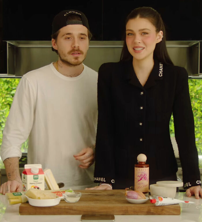 Brooklyn Beckham in a kitchen with a woman, standing behind a wooden board with breakfast ingredients arranged on it. Brooklyn Beckham in a kitchen with a woman, standing behind a wooden board with breakfast ingredients arranged on it.