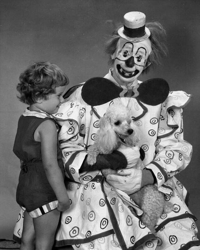 Old black and white photo of a creepy clown holding a poodle while a young child looks on with curiosity.
