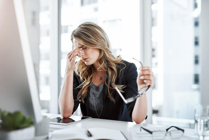 Stressed woman in office holding glasses, rubbing eyes, showing frustration with pointless and infuriating office rules.