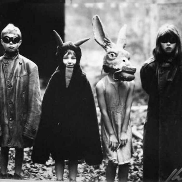 Old black and white pics showing children in eerie costumes including a large animal mask and dark cloaks in a creepy outdoor setting.