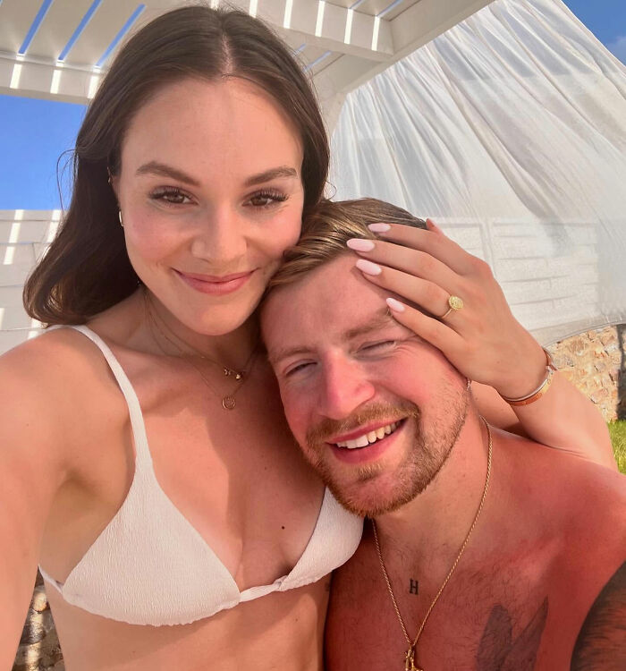 Adam Peaty smiling with woman in white bikini, embracing outdoors under bright sunlight, casual and happy moment.