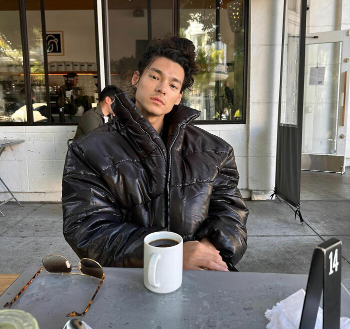 Young man in a black puffer jacket sitting at a cafe table with coffee, reflecting the SF bartender viral response story.