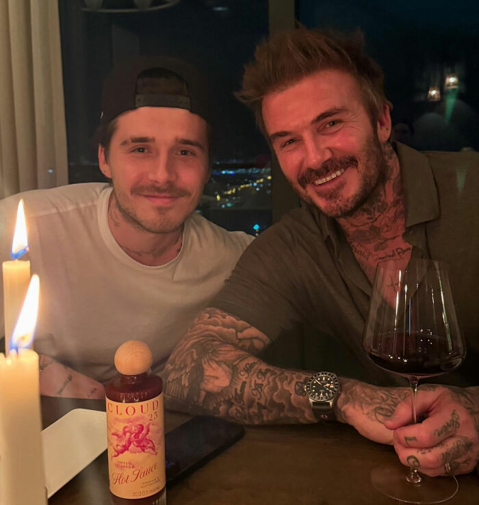 Brooklyn Beckham and David Beckham smiling at a dinner table with wine and candles amid family feud talk. Brooklyn Beckham and David Beckham smiling at a dinner table with wine and candles amid family feud talk.