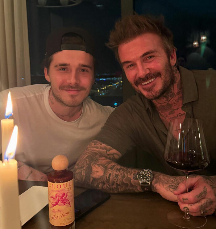 Brooklyn Beckham and a man smiling at dinner, showcasing tattoos and a bottle of hot sauce on the table. Brooklyn Beckham and a man smiling at dinner, showcasing tattoos and a bottle of hot sauce on the table.