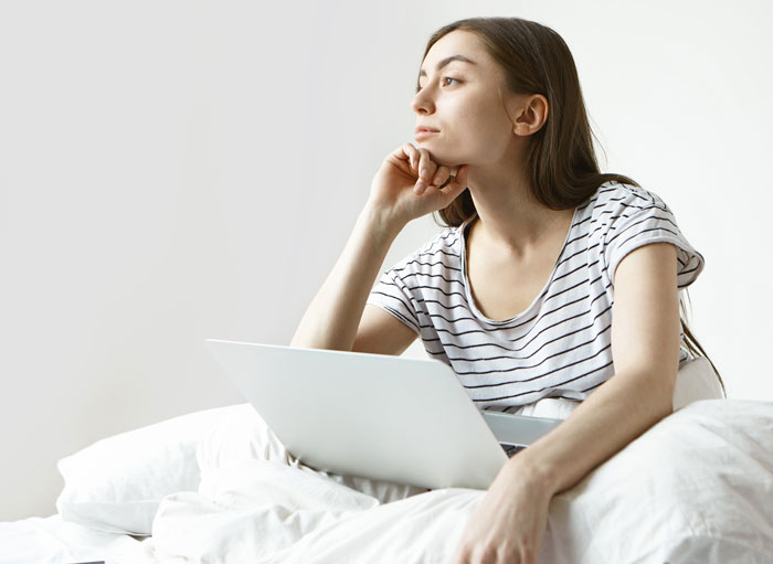 Young woman with laptop looking thoughtful while sitting on bed, relating to crazy hair stylist story and weird shrine.