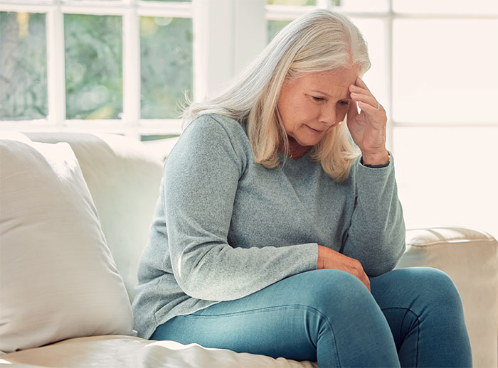 Older woman looking distressed sitting on a couch, reflecting emotions related to hiding finances from her mother-in-law. Older woman looking distressed sitting on a couch, reflecting emotions related to hiding finances from her mother-in-law.