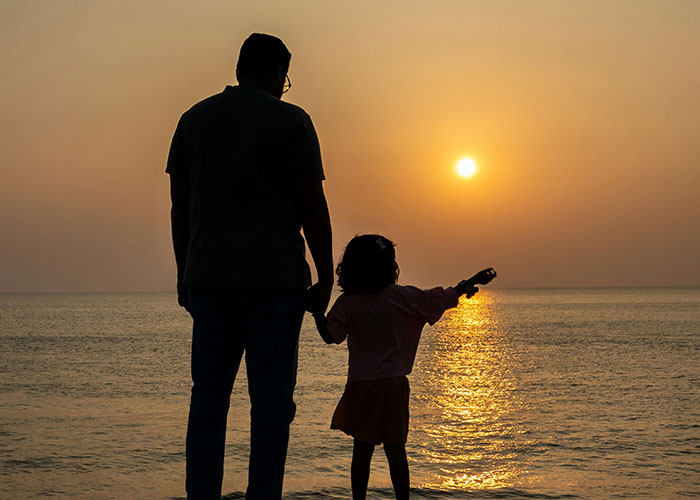 Silhouette of a man and child holding hands by the ocean at sunset, reflecting on not being the biological parent.