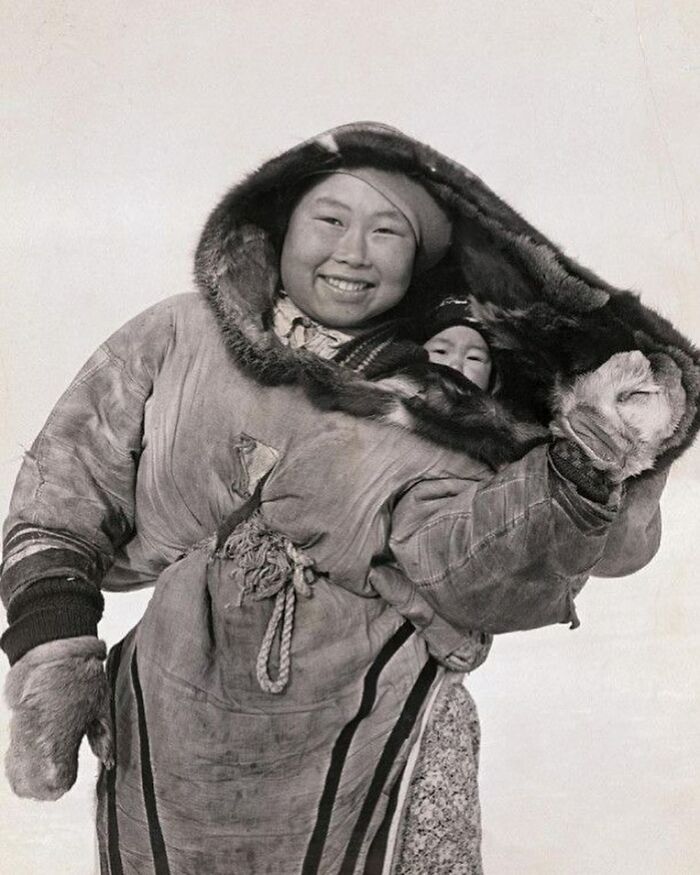 Smiling indigenous woman wearing traditional winter clothing carrying a child, a photograph showing history of humanity.