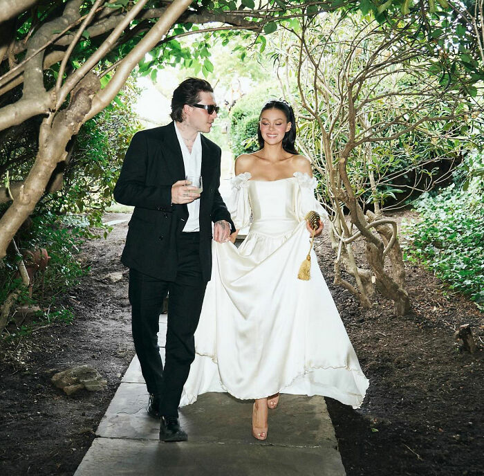 Brooklyn Beckham and Nicola Peltz walking together outdoors, dressed elegantly amid lush greenery and trees.